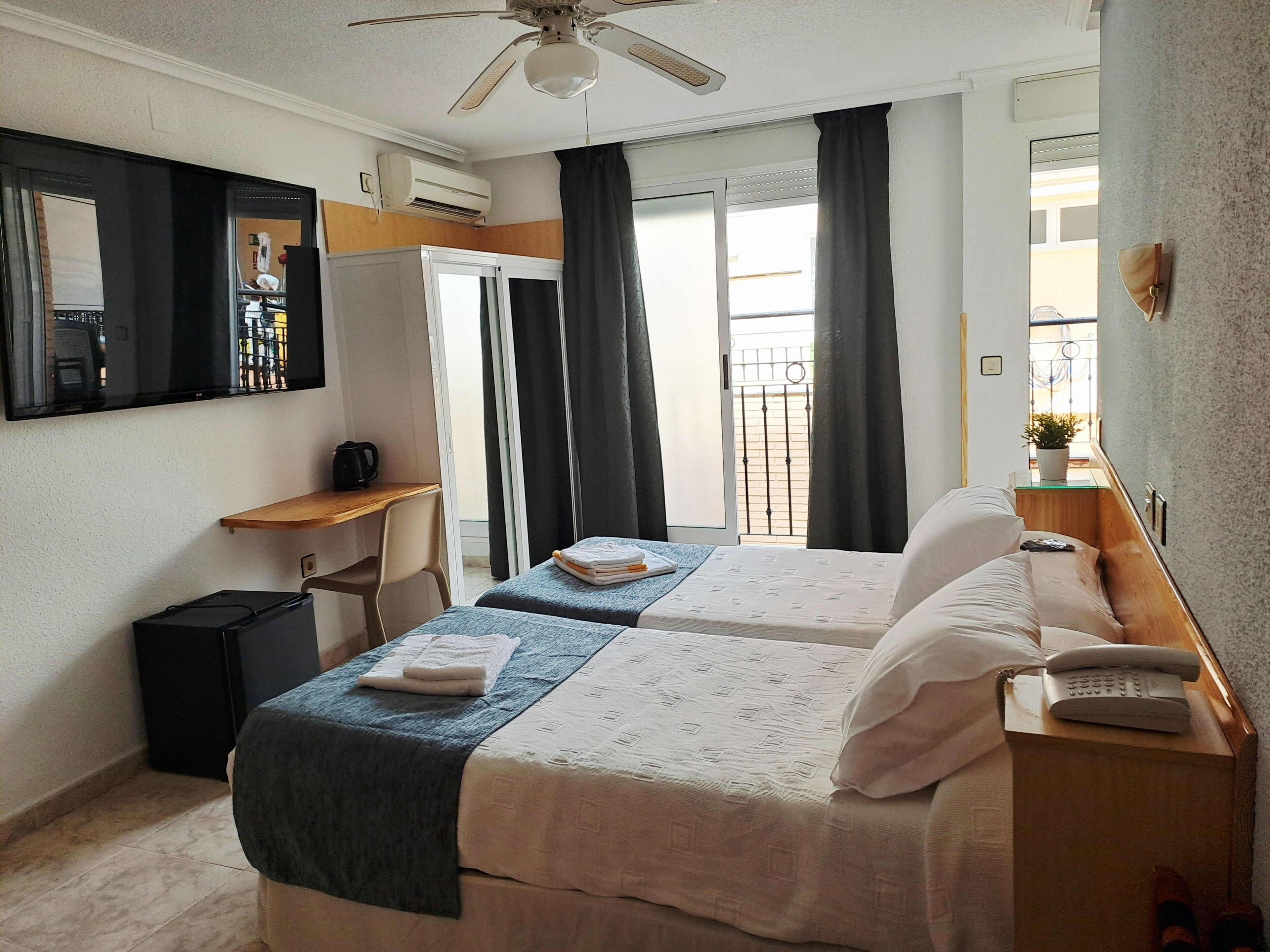 Ein Hotelzimmer mit zwei Betten, einem Schreibtisch, einem Stuhl, einem Deckenventilator, einer Klimaanlage, einem kleinen Tisch mit Pflanze und einem Fenster mit Vorhängen.