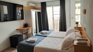 Ein Hotelzimmer mit zwei Betten, einem Schreibtisch, einem Stuhl, einem Deckenventilator, einer Klimaanlage, einem kleinen Tisch mit Pflanze und einem Fenster mit Vorhängen.