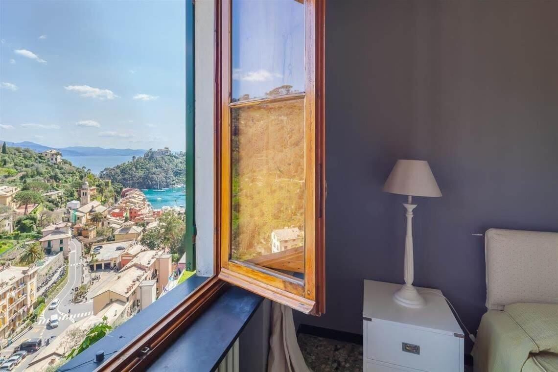 A room with a large window overlooking a coastal town, a bedside table, and a lamp.