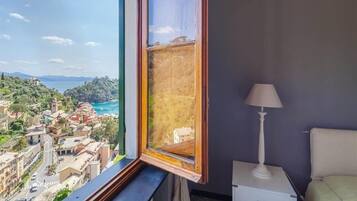 A room with a large window overlooking a coastal town, a bedside table, and a lamp.