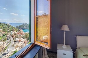 Une chambre avec une grande fenêtre donnant sur une ville côtière, une table de chevet et une lampe.