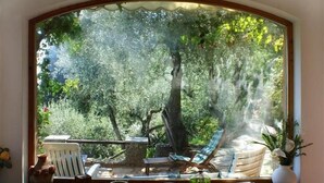 Ein Zimmer mit großem Fenster und Blick auf einen Garten. Ein Holztisch steht darin, auf dem eine Vase und eine Schüssel platziert sind, und ein Stuhl befindet sich vor dem Fenster.