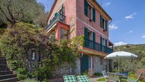 Eine pinkfarbene Villa mit grünen Fensterläden, einer Steintreppe und einer Terrasse mit zwei Liegestühlen und einem Tisch.