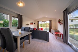 A modern living room with a dining area, a sofa, and a red bench. There are large windows with curtains, a lamp, and a view of greenery outside. - EuroParcs Brunssummerheide (Brunssum)