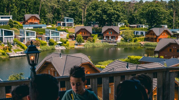 Vista para o lago com cabanas de madeira e casas modernas, uma área de refeições com pessoas e um poste de luz.