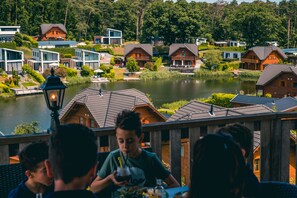 A lakeside view with wooden cabins and modern houses, a dining area with people, and a lamp post. - EuroParcs Brunssummerheide (Brunssum)