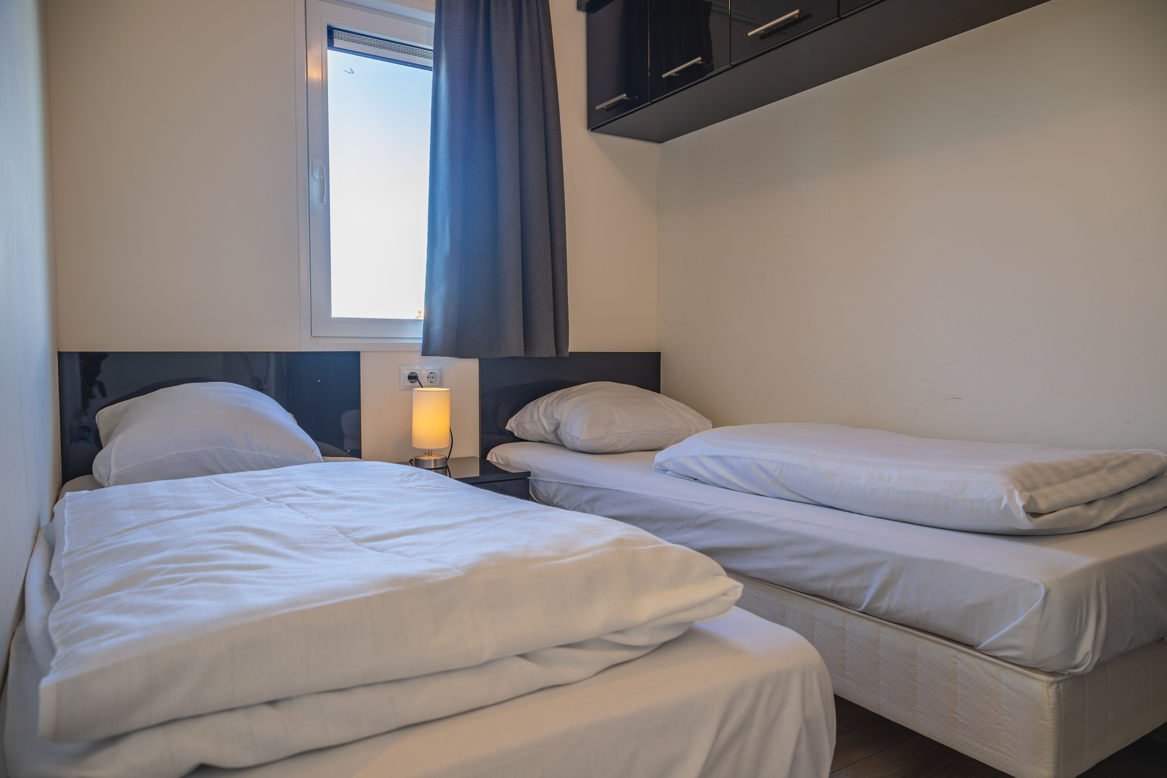Una habitación con dos camas, una ventana con cortinas, una lámpara de noche y un armario negro sobre las camas.