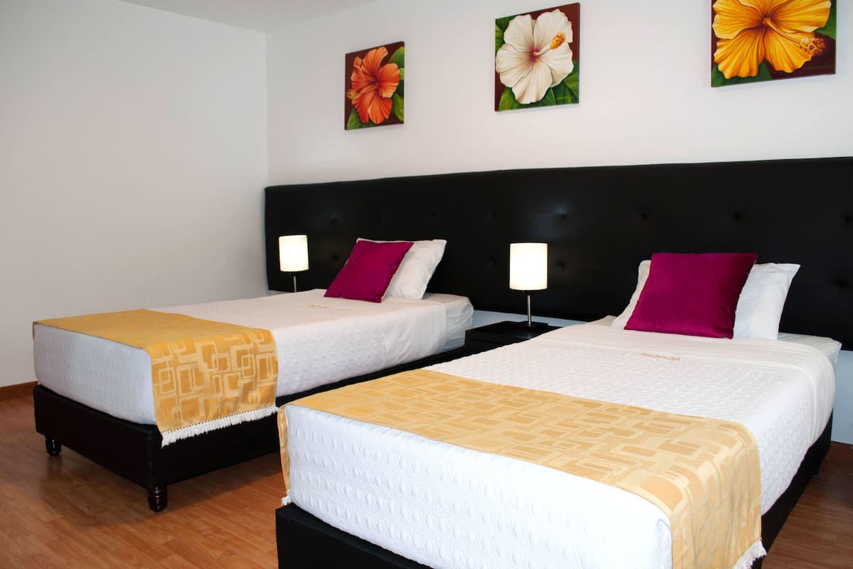 Two double beds with white linens and patterned throw blankets, a black headboard, and three floral wall art pieces.