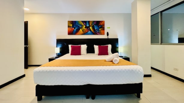 Una habitación de hotel moderna con una cama grande, un cabecero con un cuadro abstracto y colorido, una mesita de noche con una lámpara y una ventana con vistas.