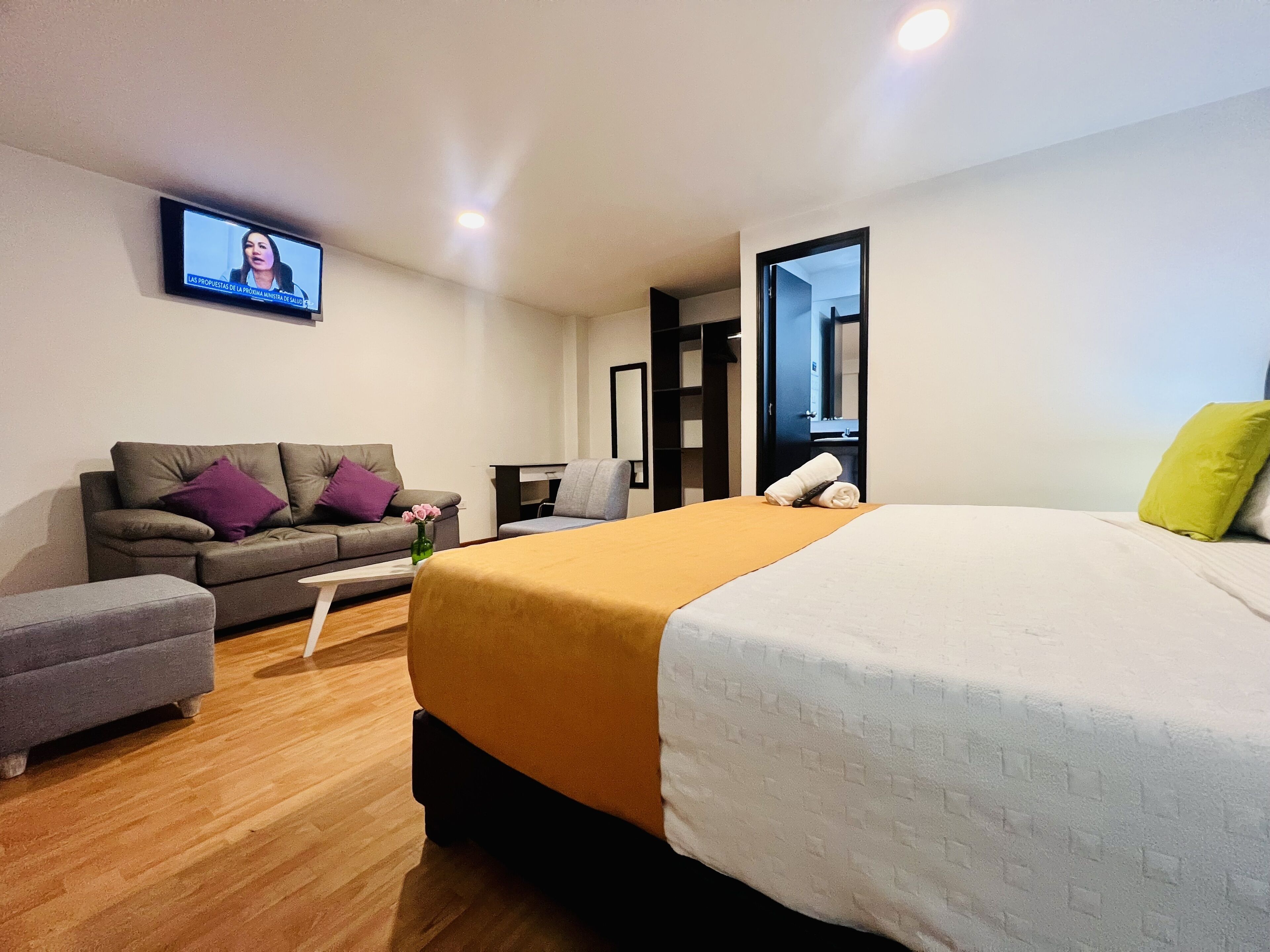 A modern hotel room with a large bed, a sofa, a small table, and a TV mounted on the wall.