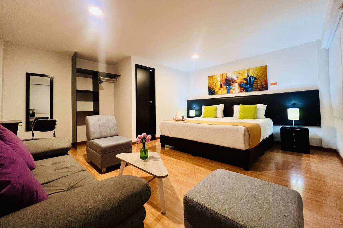 A modern hotel room with a bed, two armchairs, a small table, and a painting on the wall.