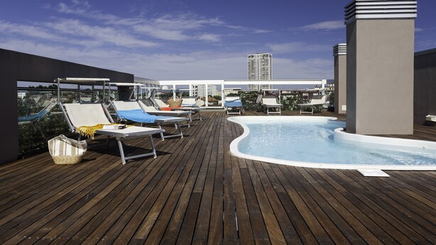 Zona de piscina en la azotea con sillones de descanso y vista a un edificio alto al fondo.