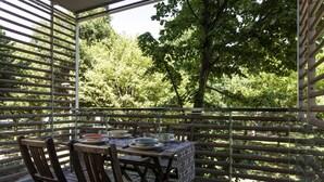 Una terraza de madera con una mesa preparada para comer, rodeada por una barandilla de madera, con vistas a un paisaje verde exuberante.
