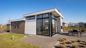 A modern two-story house with large glass windows, a flat roof, and a paved patio with outdoor furniture. - EuroParcs Veluwemeer (Nunspeet)