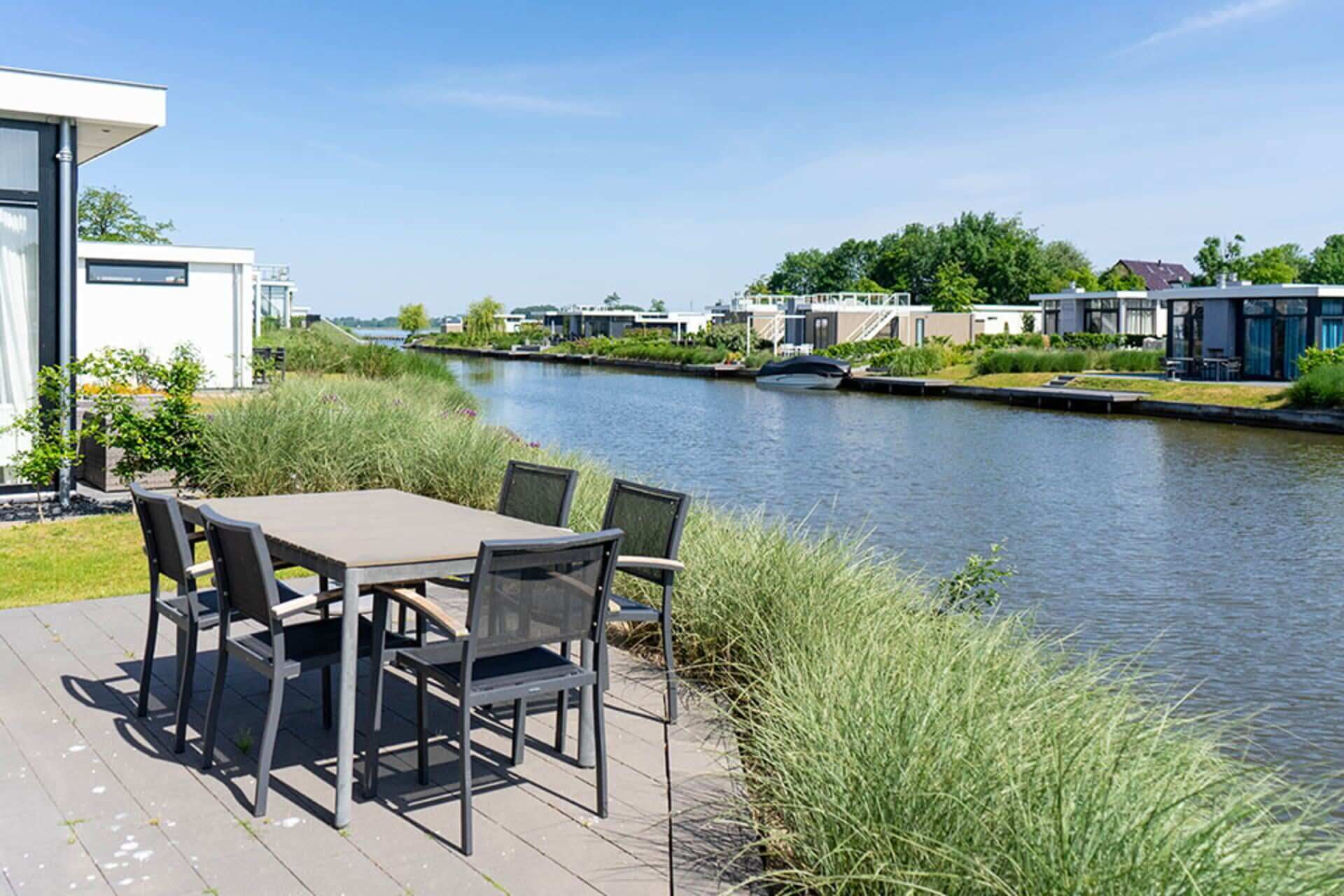 Un patio a la orilla del río con una mesa y sillas, con vistas a un canal y edificios modernos al fondo.