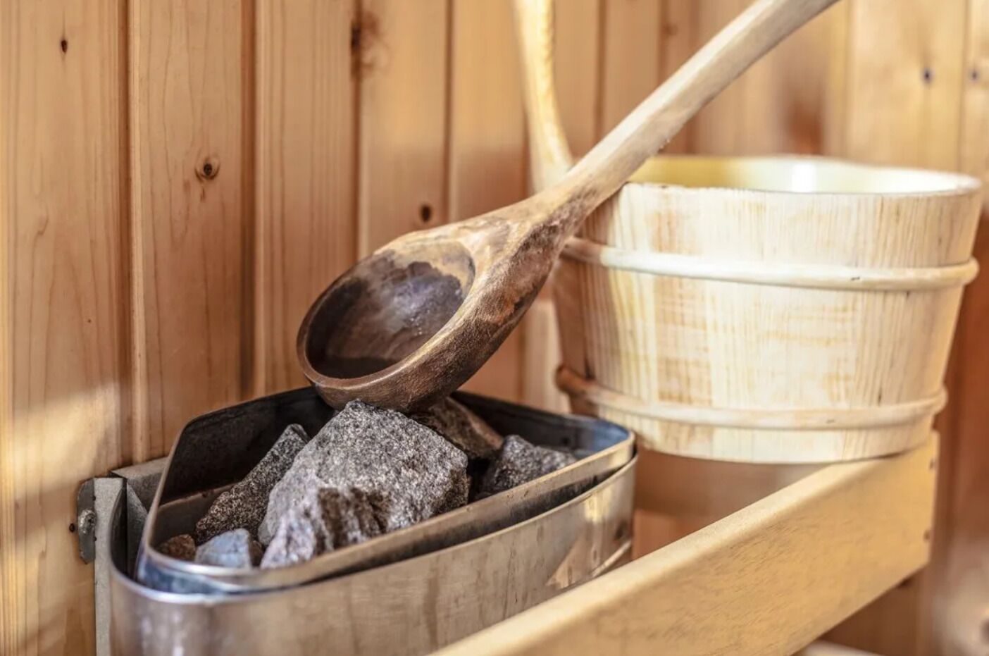 Una sauna con un banco de madera, un recipiente metálico con carbón y una cuchara de madera.