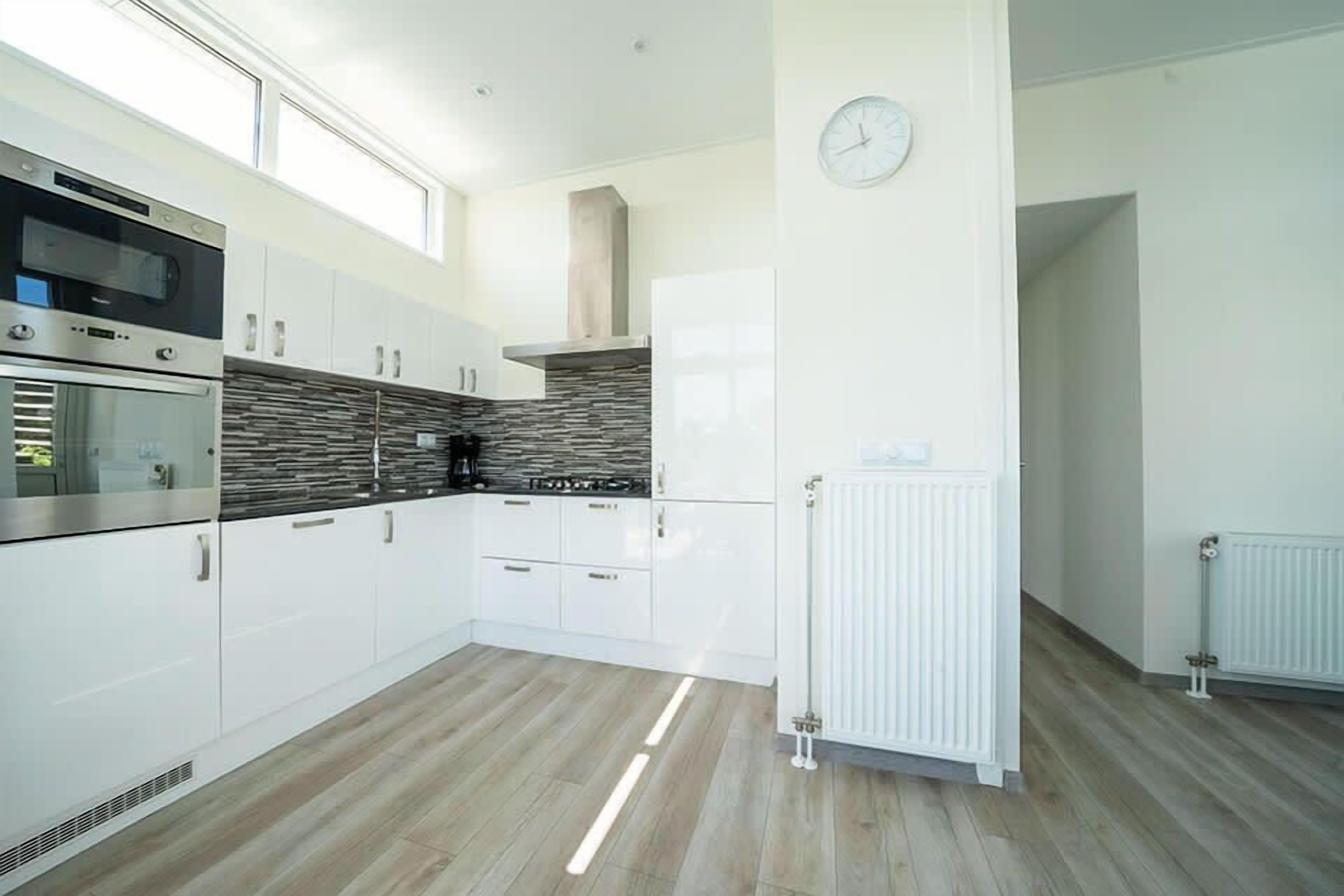 A modern kitchen with white cabinetry, a built-in oven, a microwave, a stone backsplash, and a white radiator.