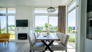 A modern living room with a dining table, comfortable seating, a fireplace, and large windows offering a view of the outdoors. - EuroParcs Veluwemeer (Nunspeet)