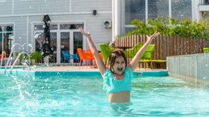 A child in a swimming pool with raised arms, surrounded by outdoor furniture and a wooden fence. - EuroParcs Veluwemeer (Nunspeet)