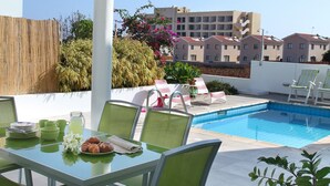 An outdoor pool area with a table and chairs, a covered seating area, and a view of surrounding buildings. - Protaras Architects Villas (Paralimni)