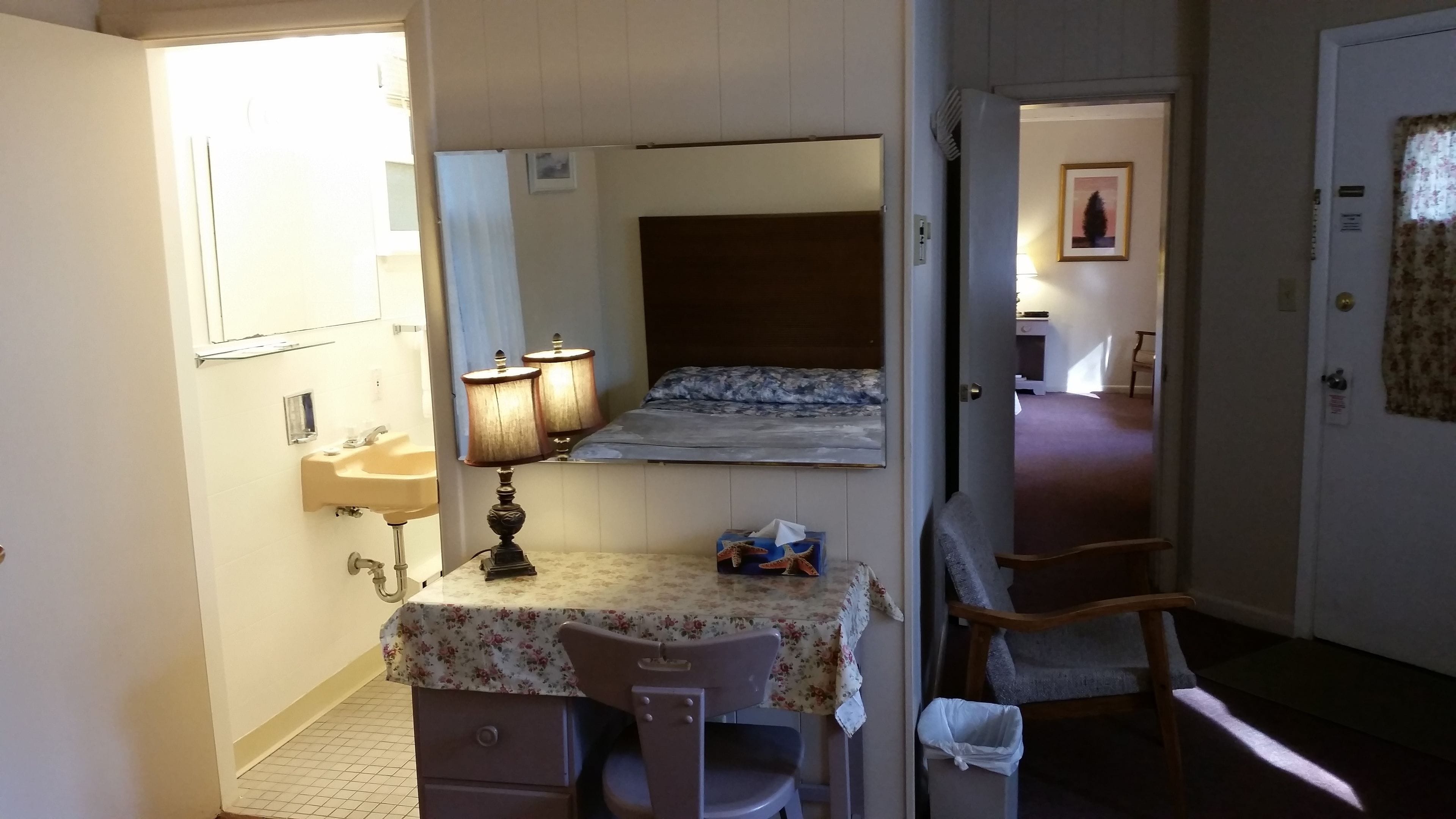 A compact hotel room with a single bed, a small desk with a chair, a bedside table with a lamp, and a bathroom with a sink and mirror.