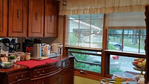 A kitchen with wooden cabinets, a chandelier, a window with blinds, and a countertop with various items. - Pine cone Motel (Penobsquis)