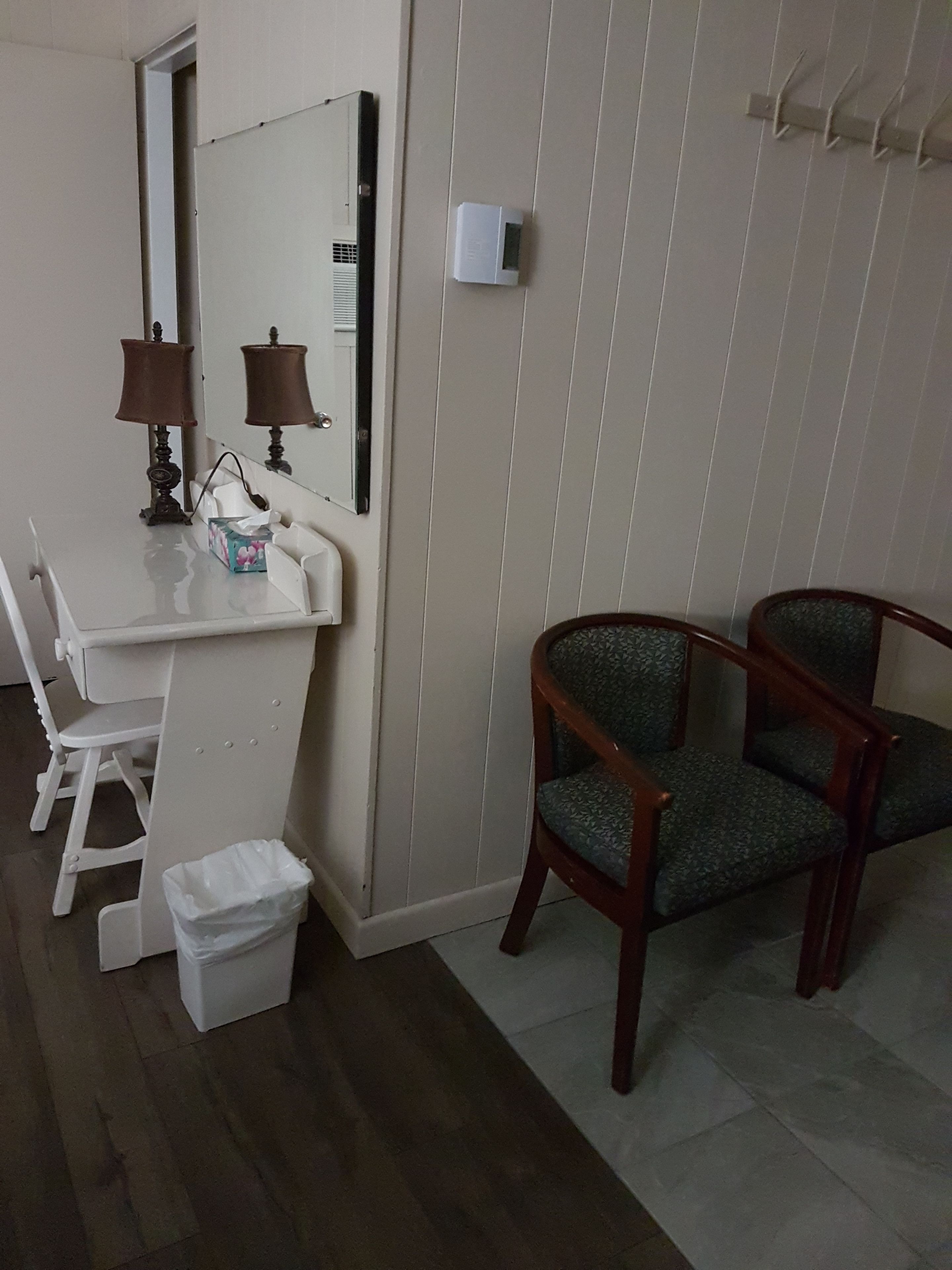 A small room with a white desk, two chairs, a mirror, a wall-mounted heater, and a trash bin.