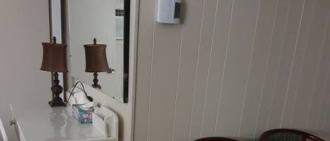 A small room with a white desk, two chairs, a mirror, a wall-mounted heater, and a trash bin.