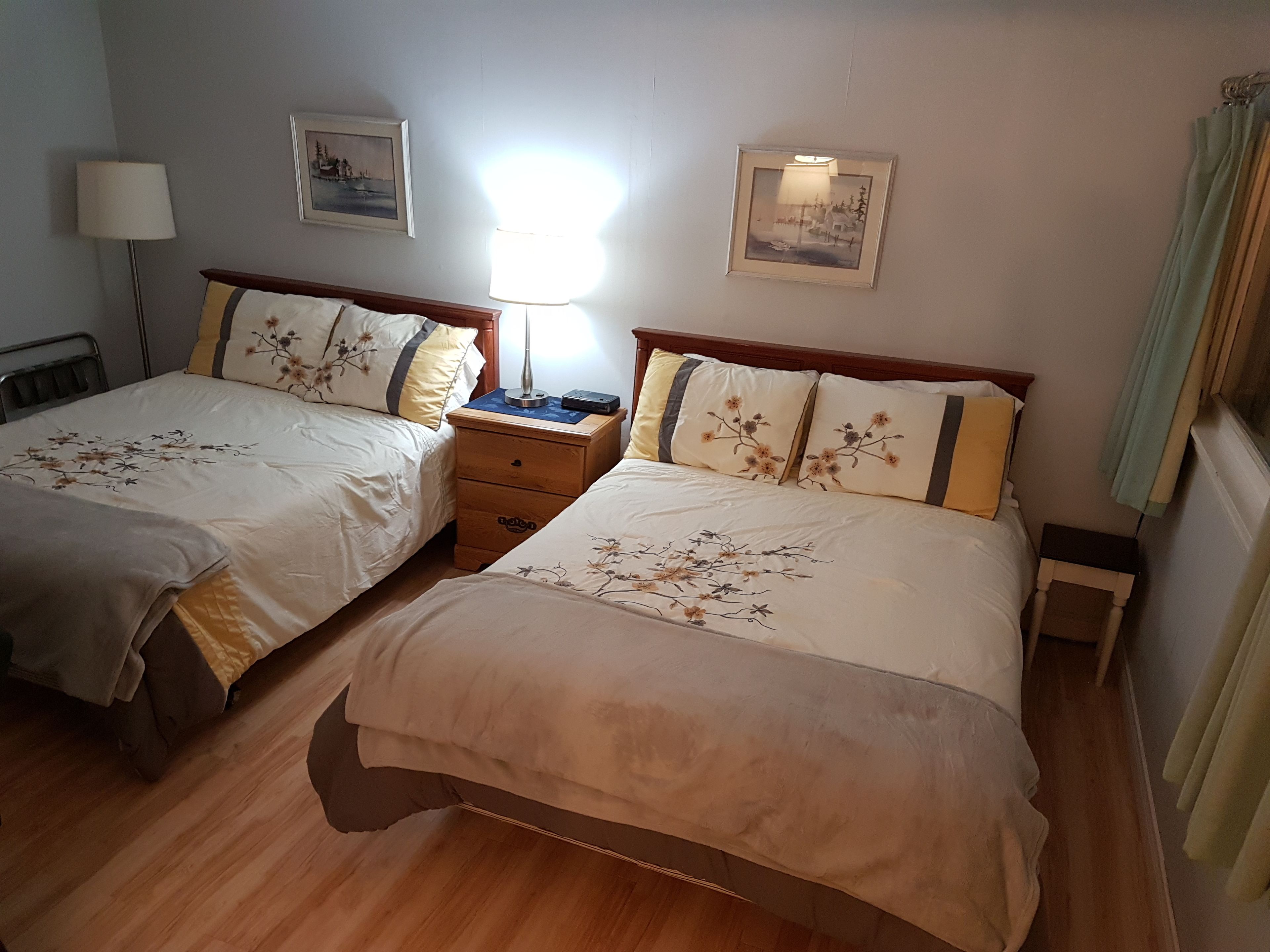 A hotel room with two beds, each with floral bedding and a wooden headboard. There are bedside tables with lamps, a small nightstand, and framed pictures on the wall.