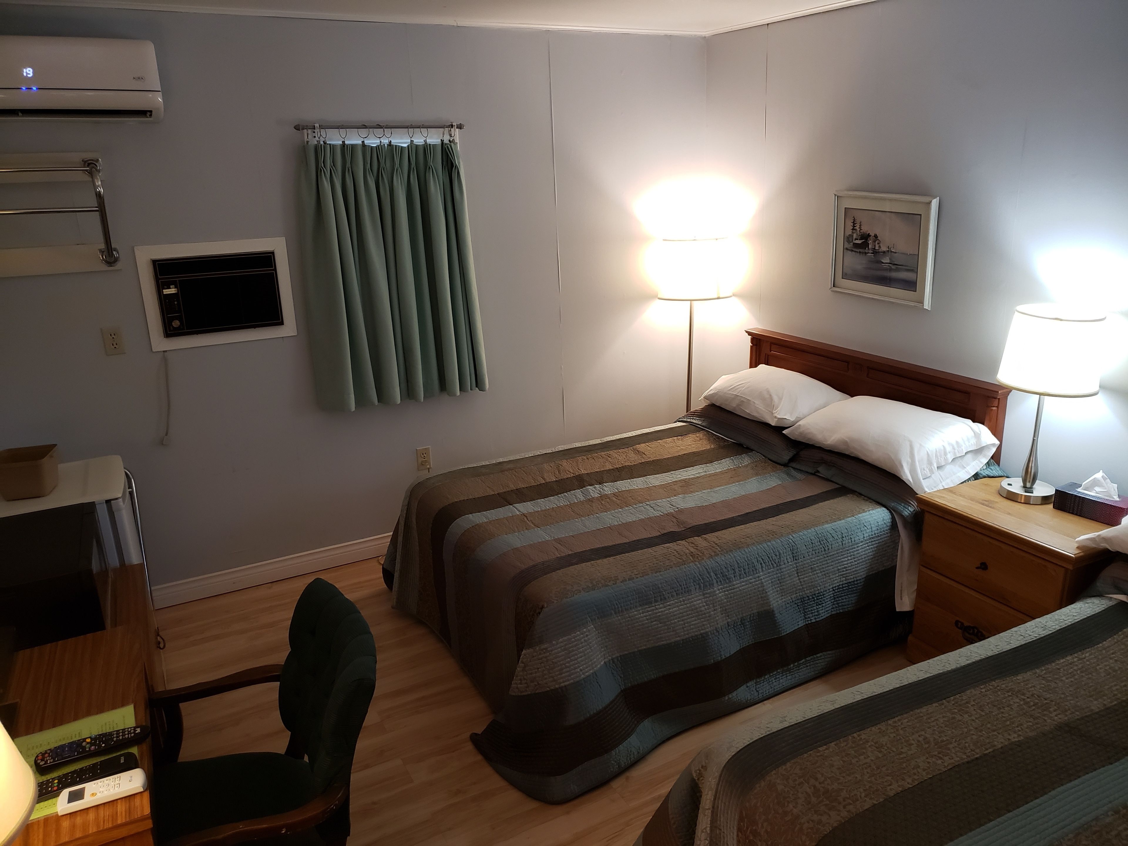 Ein Hotelzimmer mit zwei Betten, einem Schreibtisch mit Stuhl, einem Flachbildfernseher, einer Lampe und einem Bild an der Wand.