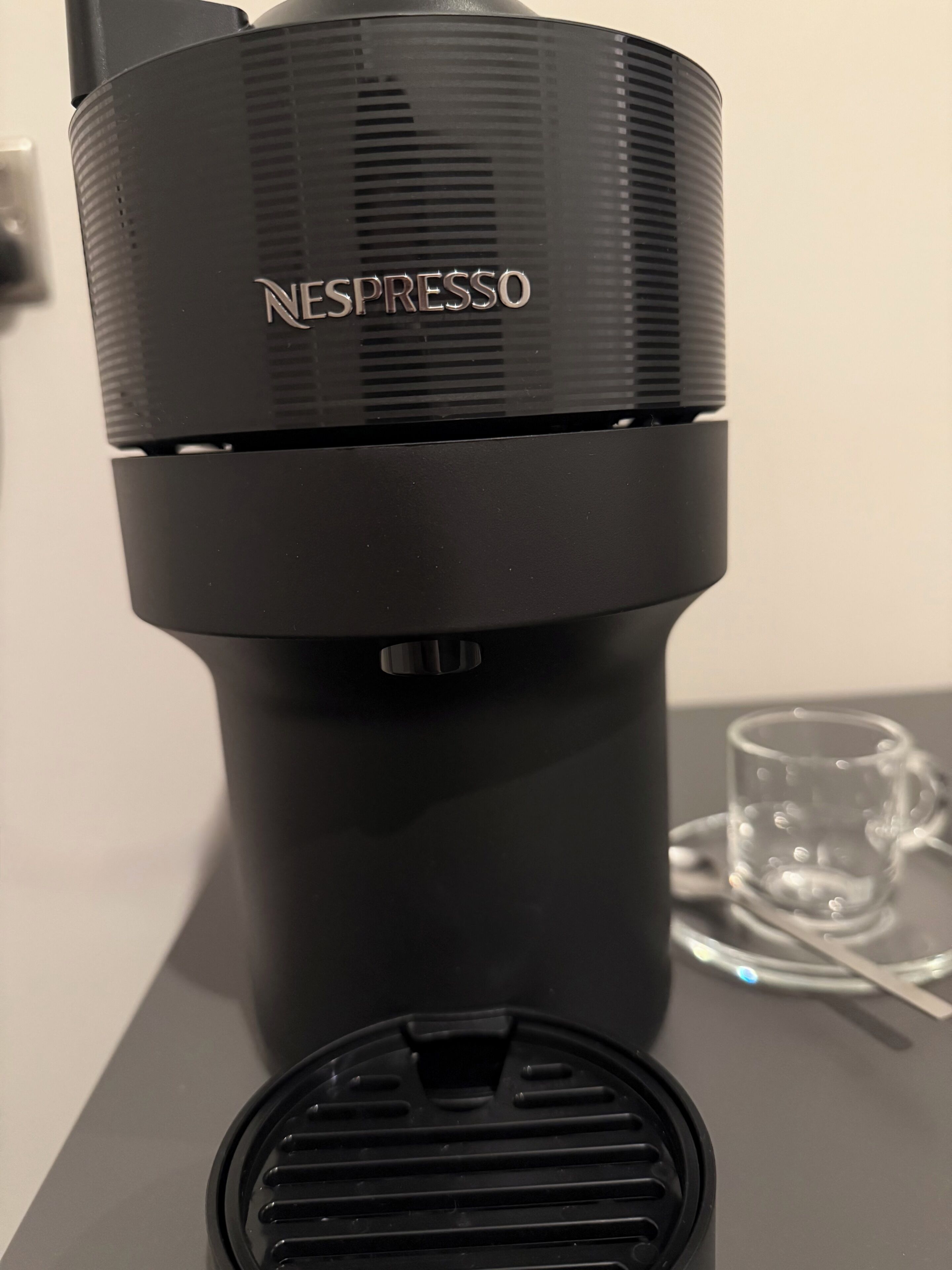 Une machine à café Nespresso avec un réservoir d’eau, un bac de récupération et une tasse en verre, posés sur une table.