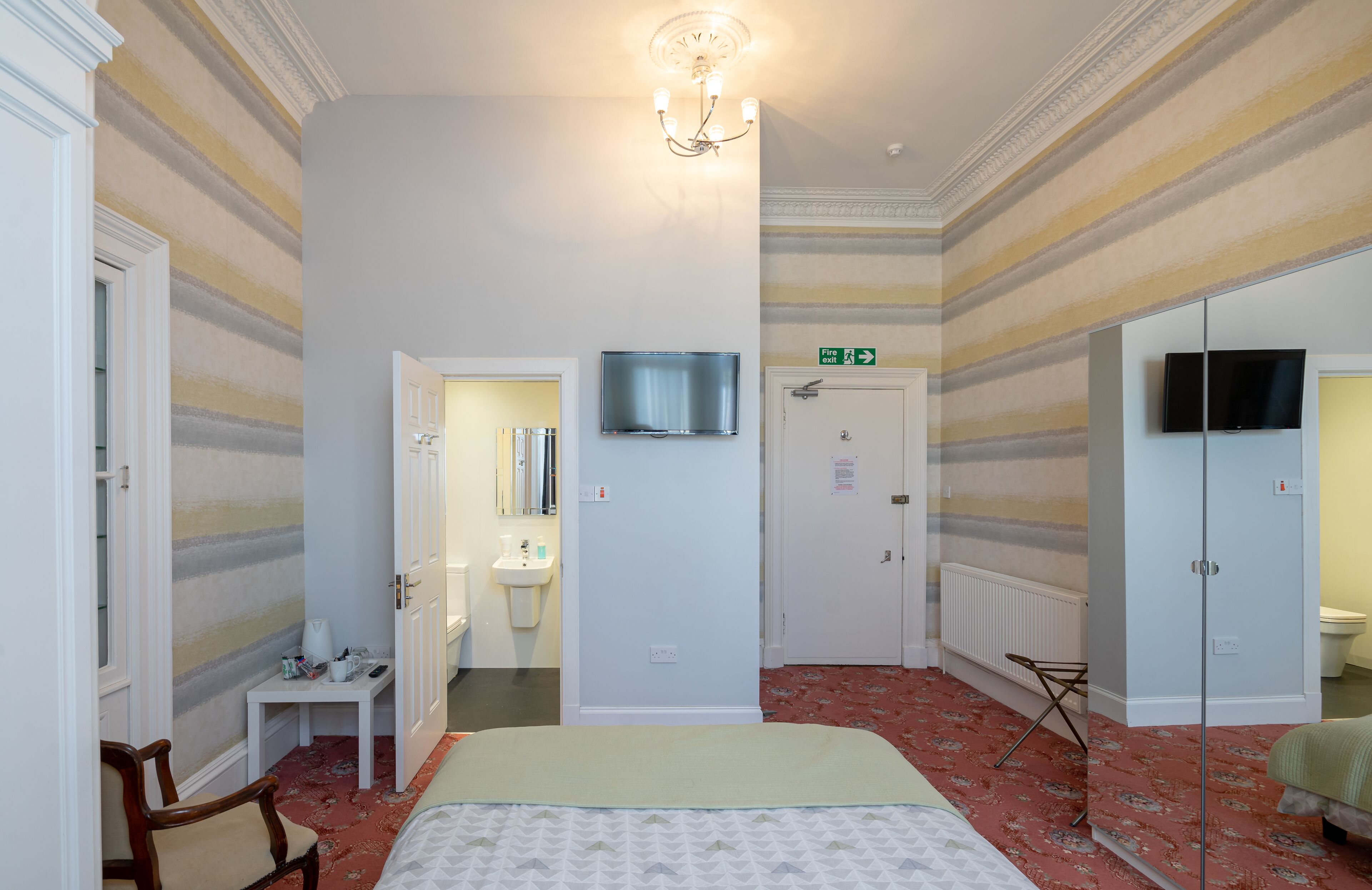 Hotelkamer met een bed, een klein tafeltje, een stoel, een televisie, een spiegel, een wastafel en een deur met een vluchtroute-bord.