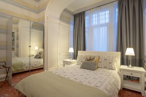 Un dormitorio de hotel con una cama grande, dos mesitas de noche con lámparas, un espejo y vistas al exterior a través de una ventana con cortinas.