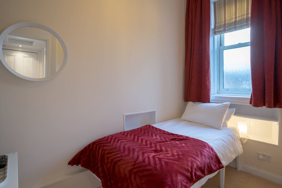 A bedroom with a bed, a round mirror, a window with curtains, and a bedside table.