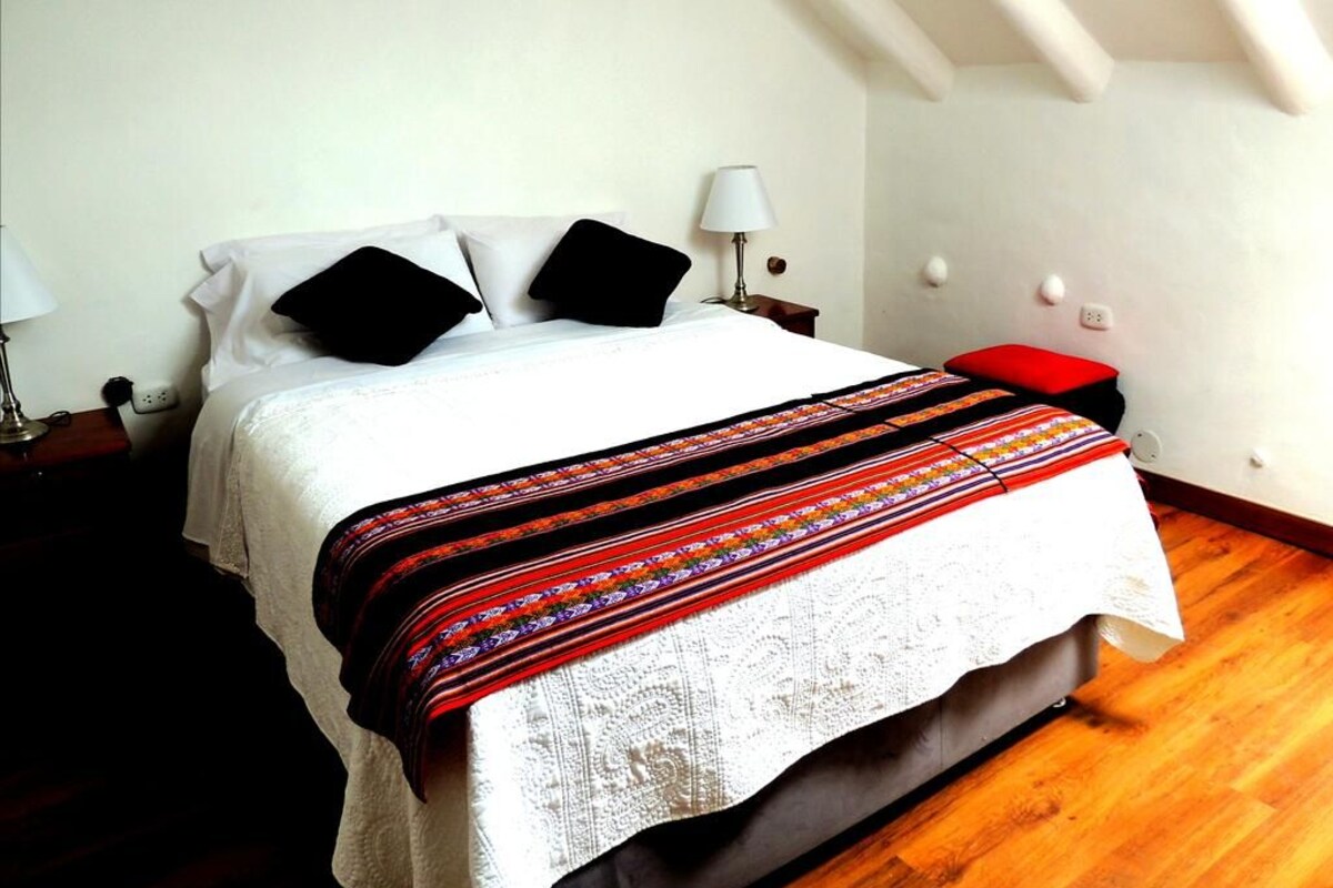 Un letto rifatto con una coperta a motivi, un comodino con una lampada e una panca rossa.