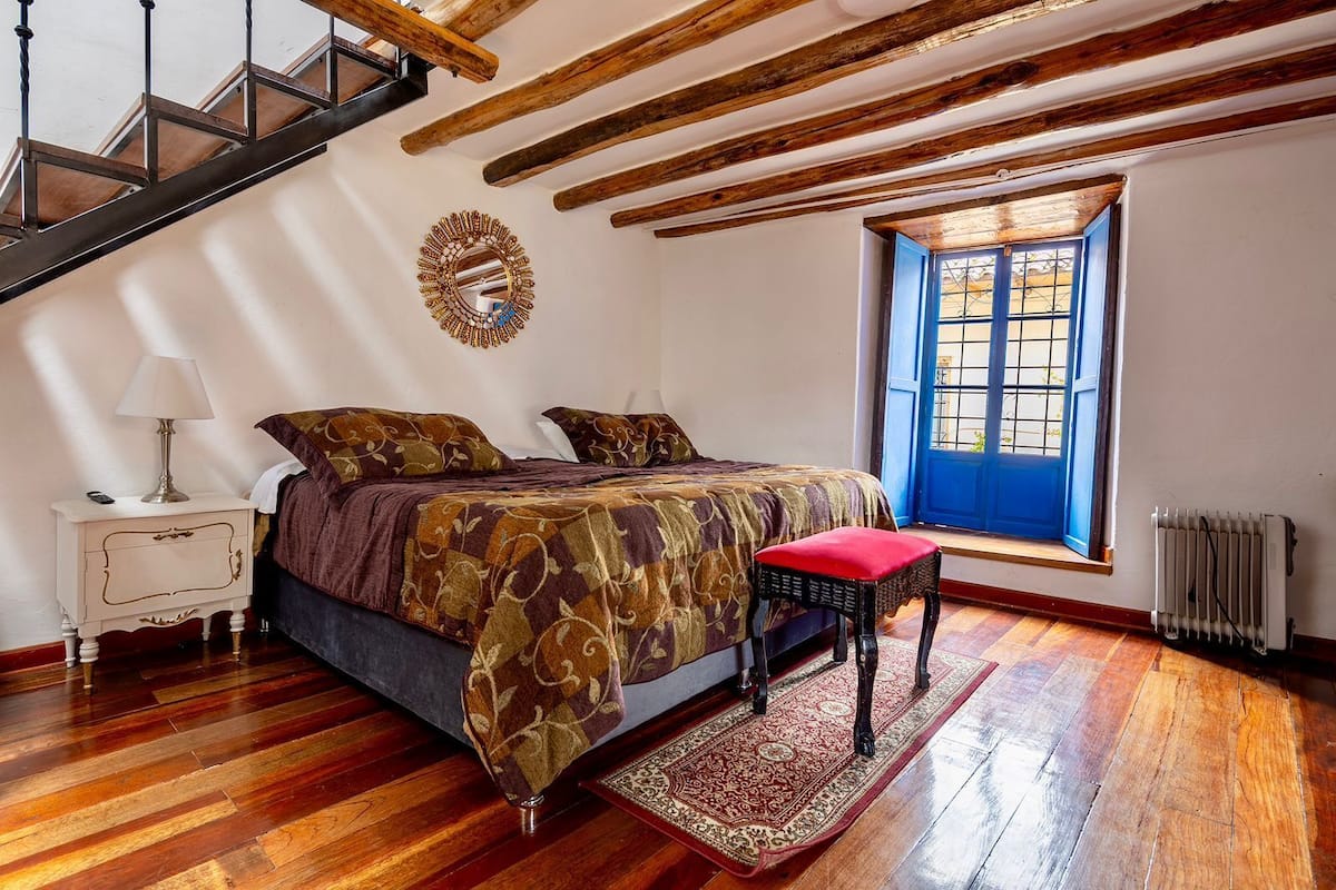 Un dormitorio con suelo de madera, una cama con ropa de cama estampada, una mesita de noche con lámpara, un banco rojo, una ventana con contraventanas azules y un techo de madera.