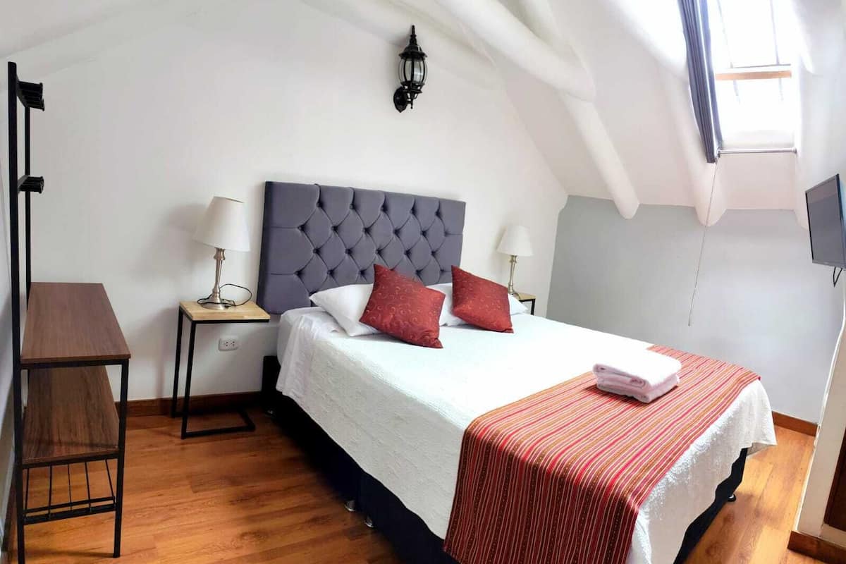Un dormitorio con cama, mesitas de noche, lámpara, televisor y ventana.