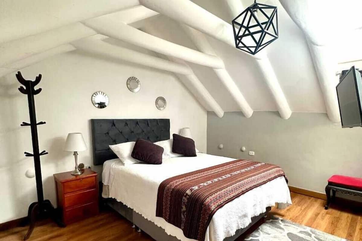 Un dormitorio con techo inclinado, una cama con cabecera oscura, una mesita de noche con lámpara y un perchero.