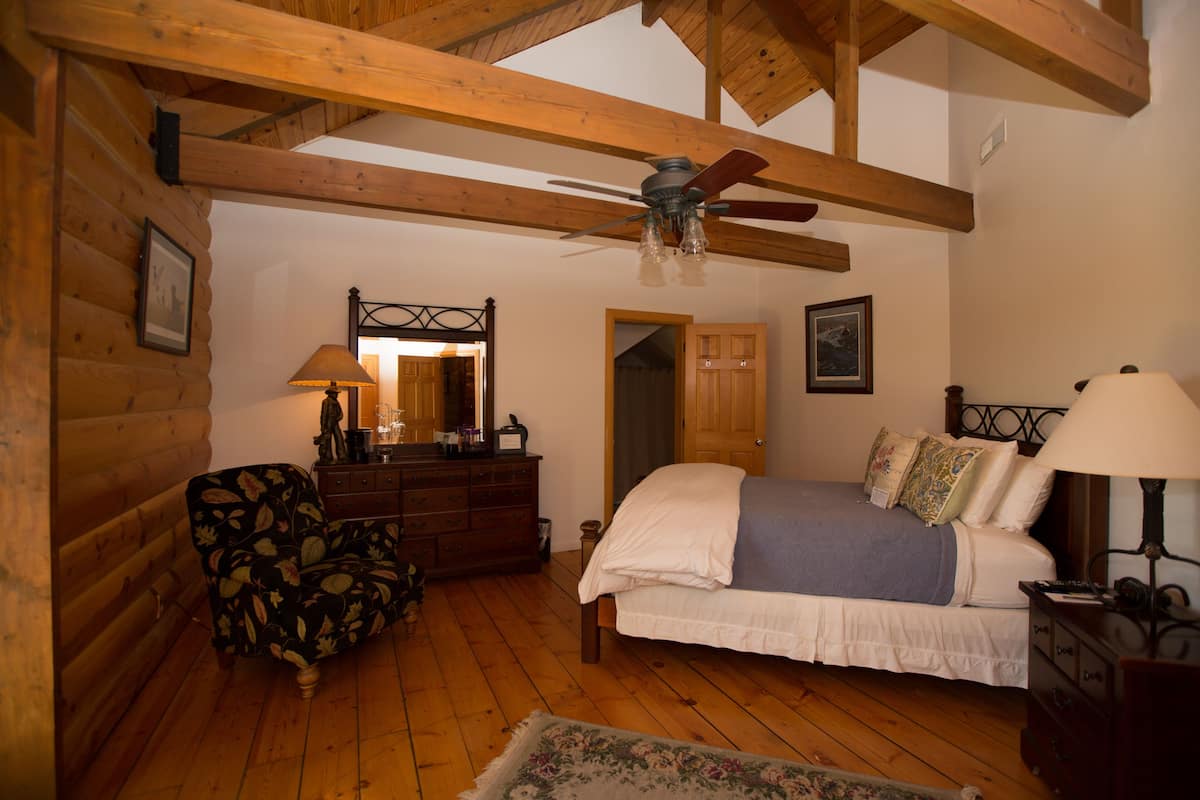 A log cabin-style bedroom with a wooden ceiling, a bed with white and blue bedding, a wooden dresser, a chair, and a mirror.