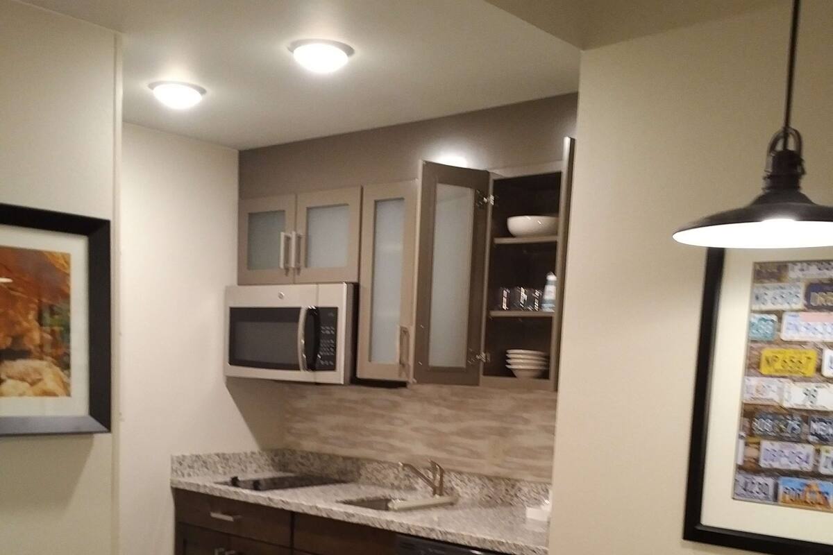 A compact kitchen with dark wooden cabinets, a microwave, and a dishwasher. There is a dining table with chairs and a framed picture on the wall.