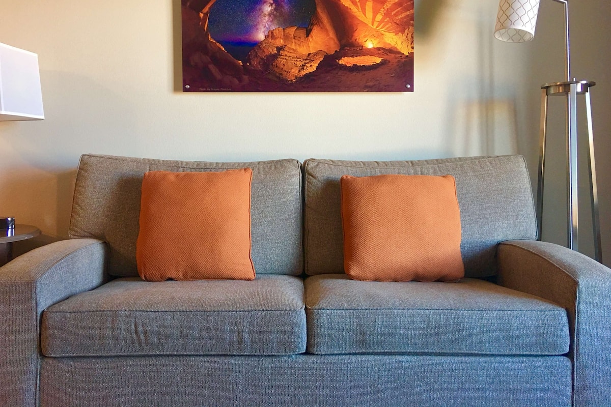 A grey sofa with two orange cushions, a framed artwork depicting a celestial scene, and a floor lamp with a patterned shade.