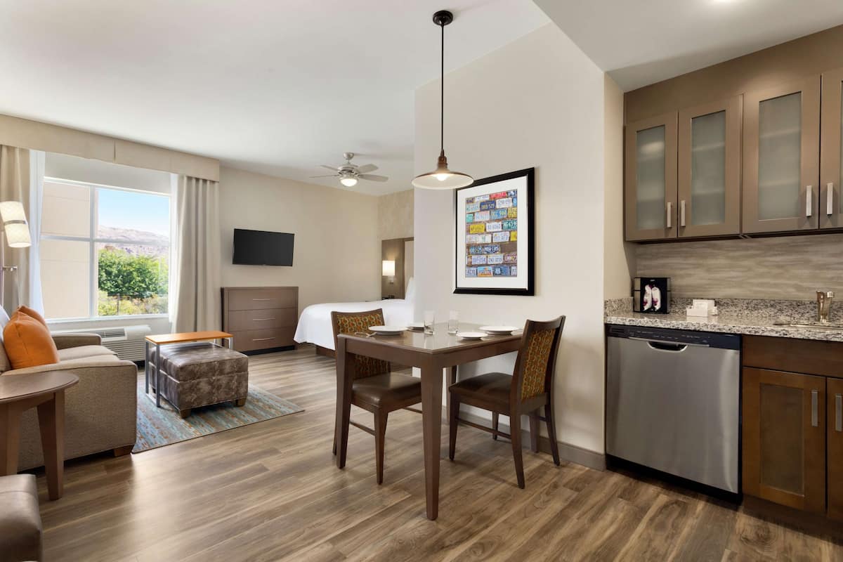 A modern hotel room with a dining area, kitchenette, and a large window offering a view of the outdoors.