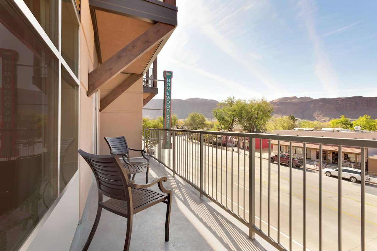 A balcony with two wooden chairs and a table, overlooking a parking lot and distant mountains.