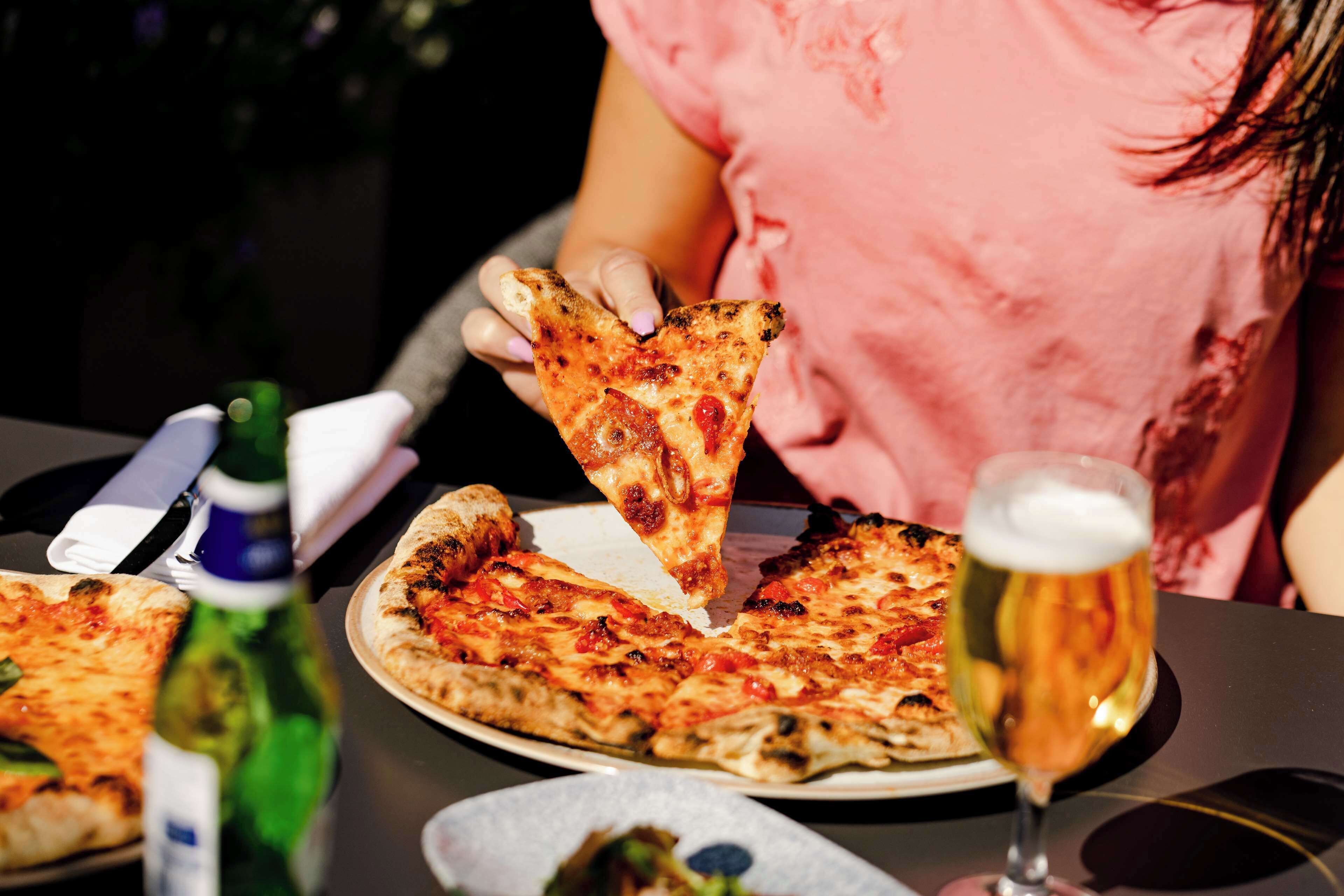 Pizza op een bord, met een plak die wordt opgetild, een glas bier en een fles olijfolie op een tafel.