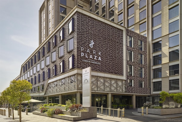 A modern building with a sign reading "Park Plaza London Waterloo." It features a brick facade, multiple windows, and a landscaped area with plants and a tree.