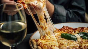 Una persona sosteniendo una porción de pizza con queso derretido, una copa de vino blanco y un plato de papas fritas sobre una mesa.