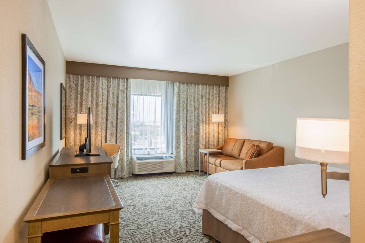 A hotel room with a bed, a desk, a chair, a sofa, and a window with curtains.