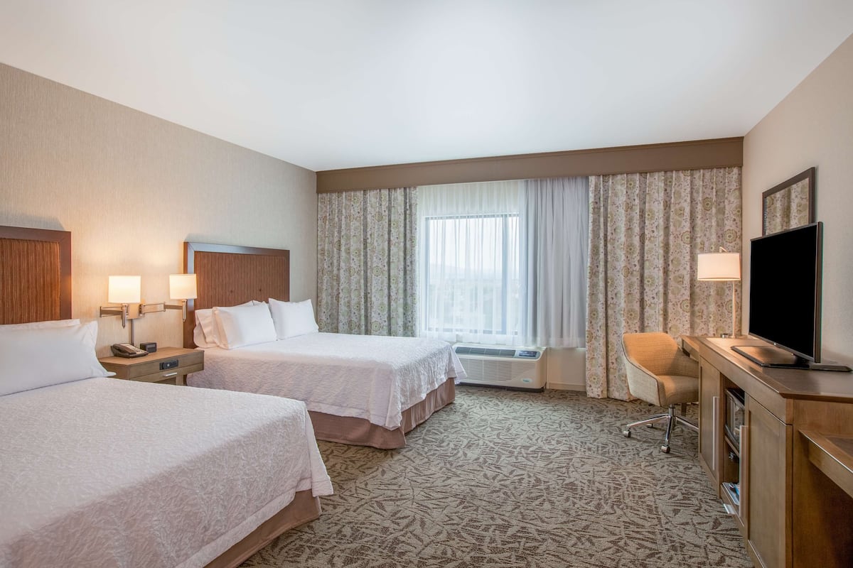 A hotel room with two beds, a desk with a television, a chair, and a window with curtains.