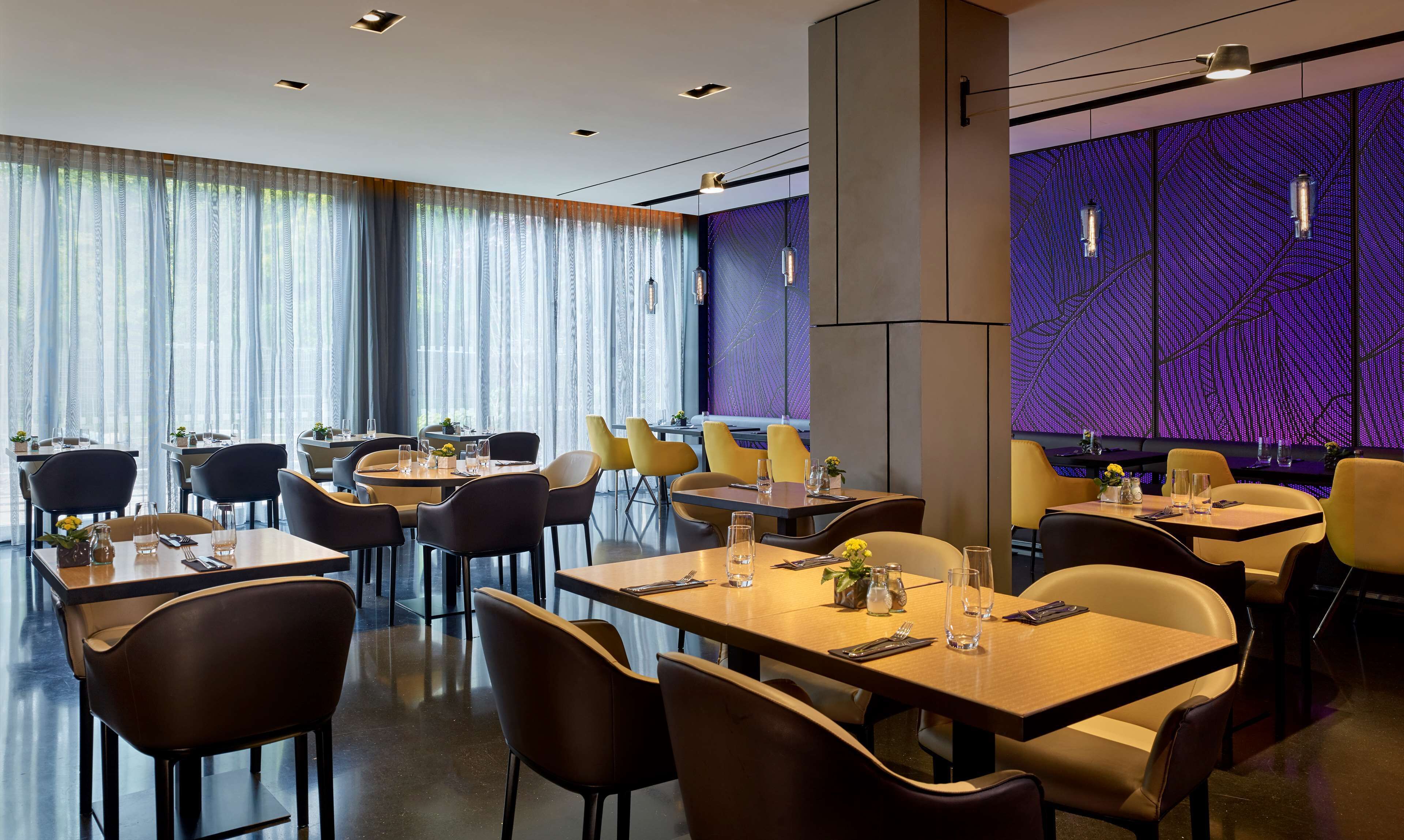 Restaurante moderno com mesas redondas e cadeiras amarelas, janelas amplas com cortinas leves e uma parede com padrão roxo.