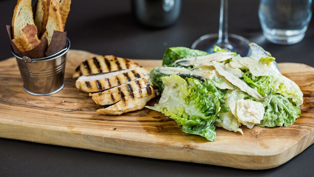 Una tabla de madera con pollo a la parrilla, ensalada y palitos de pan.
